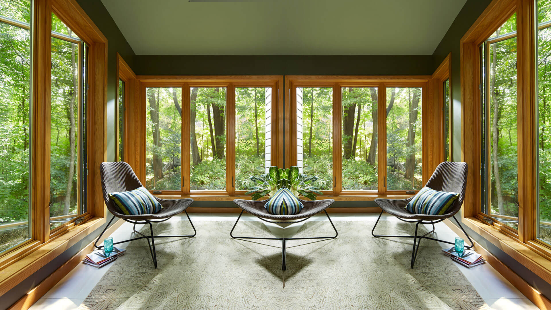 Rustic-style sunroom with two chairs, a coffee table, and large woodgrain casement windows overlooking forest.